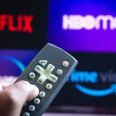 Dłoń trzymająca pilot do telewizora na tle ekranu z logo HBO Max, Netflix i Prime Video.